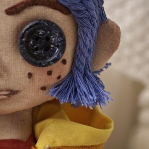 K&ouml;nnte beinhalten: Handgefertigte Puppe mit blauen Garnhaaren, einem gro&szlig;en schwarzen Knopfauge und gestickten Sommersprossen. Die Puppe tr&auml;gt ein gelbes und rotes Outfit mit einem detaillierten, gen&auml;hten Gesicht. Das Haar der Puppe ist in einem geflochtenen Design gestaltet.