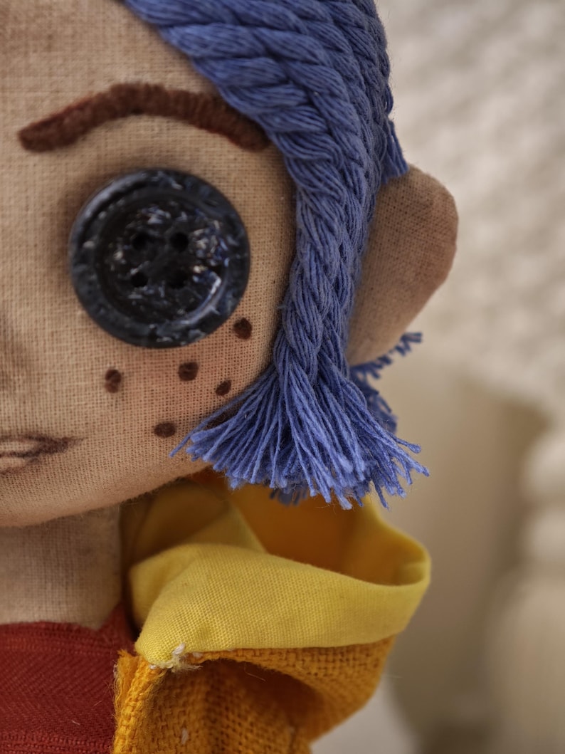 K&ouml;nnte beinhalten: Handgefertigte Puppe mit blauen Garnhaaren, einem gro&szlig;en schwarzen Knopfauge und gestickten Sommersprossen. Die Puppe tr&auml;gt ein gelbes und rotes Outfit mit einem detaillierten, gen&auml;hten Gesicht. Das Haar der Puppe ist in einem geflochtenen Design gestaltet.