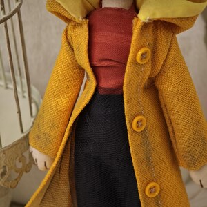 K&ouml;nnte beinhalten: Handgefertigte Puppe mit einem senfgelben Mantel mit gelber Kapuze und drei Kn&ouml;pfen. Die Puppe hat blaue Haare, ein rotes Oberteil und einen schwarzen Rock. Das Gesicht der Puppe ist handgen&auml;ht.