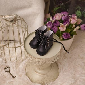 Może przedstawiać: Para miniaturowych czarnych skórzanych butów z białymi szwami i sznurowadłami. Buty są prezentowane na małym, ozdobnym, kremowym cokole. W tle dekoracyjna klatka dla ptaków i bukiet fioletowych i różowych kwiatów.