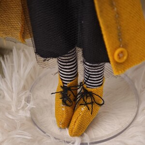 K&ouml;nnte beinhalten: Nahaufnahme der Beine und F&uuml;&szlig;e einer Puppe. Die Puppe tr&auml;gt schwarz-wei&szlig; gestreifte Socken, gelbe Schuhe mit schwarzen Schn&uuml;rsenkeln und einen gelben Mantel mit gro&szlig;en Kn&ouml;pfen. Die Puppe steht auf einem transparenten, kreisf&ouml;rmigen St&auml;nder.