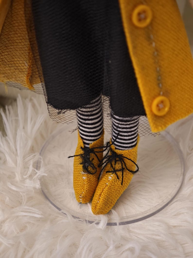 K&ouml;nnte beinhalten: Nahaufnahme der Beine und F&uuml;&szlig;e einer Puppe. Die Puppe tr&auml;gt schwarz-wei&szlig; gestreifte Socken, gelbe Schuhe mit schwarzen Schn&uuml;rsenkeln und einen gelben Mantel mit gro&szlig;en Kn&ouml;pfen. Die Puppe steht auf einem transparenten, kreisf&ouml;rmigen St&auml;nder.