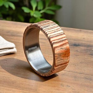 May include: A wooden ring with a striped pattern in shades of brown and tan. The interior of the ring is a silver metal. The ring is sitting on a wooden surface.