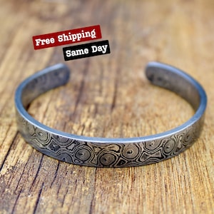 Puede incluir: Brazalete plateado con un patrón arremolinado único. El brazalete abierto presenta un diseño detallado y texturizado en tonos grises y negros. El brazalete se muestra sobre una superficie de madera.