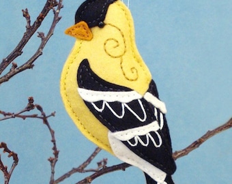 May include: A felt ornament of a yellow bird with black and white markings. The bird is perched on a branch and has a small orange beak. The ornament is handmade and has a whimsical design.