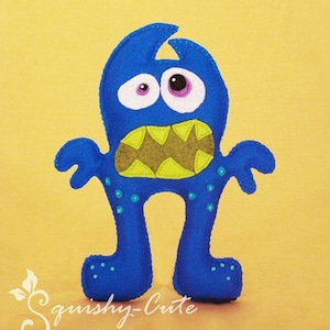 Puede incluir: Monstruo de fieltro azul con una gran sonrisa de dientes, dientes verdes y ojos saltones. El monstruo tiene brazos y piernas de fieltro azul con puntos turquesa.
