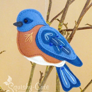 May include: Bluebird felt ornament with blue, brown, and white felt. The bird has a beaded wing and a white belly. The ornament is hanging from a branch.