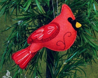 May include: A red felt cardinal bird ornament with black eyes and a yellow beak. The ornament is hanging from a white string and is surrounded by green pine branches.