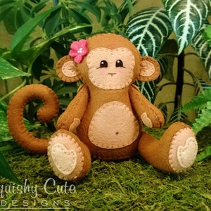 May include: A brown felt monkey toy with a pink flower on its head. The monkey has a curled tail and is sitting on a green mossy surface.