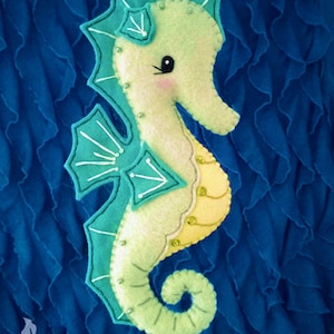 May include: A green and blue felt seahorse with a yellow belly and white eyes. The seahorse is decorated with white stitching and beads. The seahorse is on a blue background.