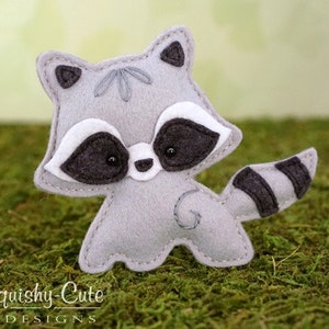 May include: A gray felt raccoon with black eyes and a black and white striped tail. The raccoon is standing on a bed of green moss.