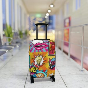 Puede incluir: Una maleta con ruedas con un colorido diseño pop art. El diseño incluye imágenes de labios, una bombilla, una cereza y la palabra "LOVE". La maleta tiene un asa y ruedas negras.