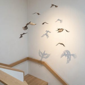 Op de afbeelding: Metalen vogelsculpturen hangen aan het plafond en werpen schaduwen op een witte muur. De vogels zijn in vlucht, met gedetailleerde vleugelontwerpen. Een houten trap met leuning is ook zichtbaar.