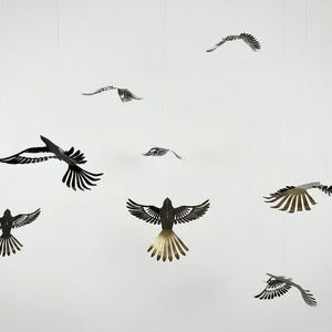 Puede incluir: Una colección de esculturas de pájaros de metal en varias etapas de vuelo, suspendidas sobre un fondo blanco. Los pájaros son principalmente negros y plateados, con diseños detallados en las alas. La disposición crea una sensación dinámica y aireada.
