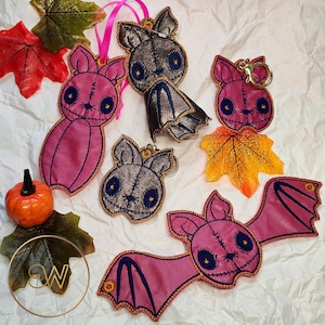 May include: A collection of bat-shaped ornaments and keychains in pink and metallic bronze faux leather with stitched details, dark blue circular eyes, and embroidered features, accompanied by artificial autumn leaves and a small orange pumpkin.