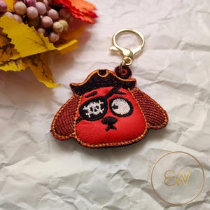 May include: A red embroidered keychain shaped like a dog head wearing a black pirate hat. The dog has a black eye patch with a white skull and crossbones design, a single white eye, and a gold-toned metal lobster clasp attached at the top.