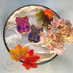 May include: Two embroidered keychains shaped like animal heads, one in bright pink and one in dark bronze, both featuring blue circular eyes and stitched details. They are displayed alongside artificial autumn leaves and a small orange pumpkin.