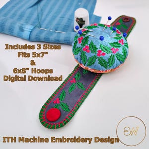 May include: A wrist pincushion with a light blue fabric top featuring embroidered green holly leaves and red berries, attached to a dark grey strap with a red button. Text reads: Includes 3 Sizes Fits 5x7" & 6x8" Hoops Digital Download ITH Machine Embroidery Design.