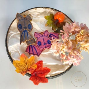 May include: Two bat-shaped keychains, one in brown and one in pink, featuring embroidered details and blue eyes. They are displayed in a round container with white tissue paper, artificial autumn leaves, a small orange pumpkin, and dried flowers.