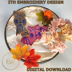 May include: Two bat-shaped embroidery designs, one in brown and one in pink, featuring blue eyes and stitched details. The text ITH EMBROIDERY DESIGN and DIGITAL DOWNLOAD is visible. The bats are displayed with artificial autumn leaves and flowers.