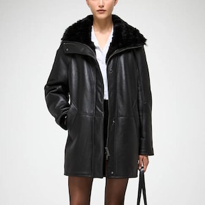Puede incluir: Un abrigo de cuero negro con cuello de piel sintética y un bolso tote negro a juego. El abrigo tiene cierre de botones y un corte entallado. La modelo lleva una camisa blanca y una falda negra. Los zapatos son tacones negros.