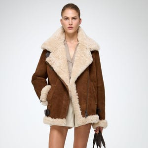Könnte beinhalten: Braune Shearling-Jacke aus Wildleder mit cremefarbenem Shearling-Futter und -Besatz. Die Jacke hat einen breiten Kragen, einen diagonalen Reißverschluss und Schnallendetails. Das Model trägt außerdem kniehohe Stiefel aus braunem Wildleder und eine braune Wildleder-Tragetasche.