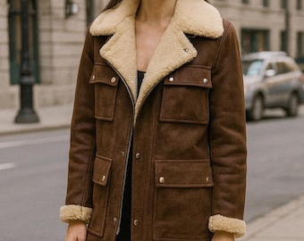 May include: A brown suede coat with a cream-colored shearling collar and cuffs. The coat features four front pockets, a zipper closure, and button details. The model is standing on a city street.
