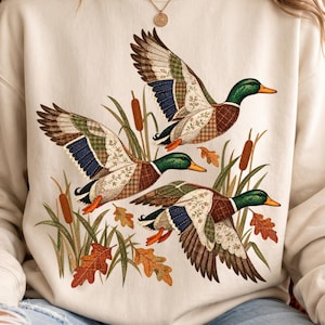 Könnte beinhalten: Ein cremefarbenes Sweatshirt mit Rundhalsausschnitt und einem Grafikdruck von drei fliegenden Stockenten. Die Flügel der Enten sind mit einem Patchwork-Muster aus verschiedenen Stoffen gestaltet, darunter Blumen- und Karomuster, umgeben von Rohrkolben.