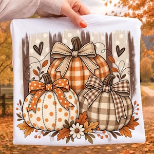 May include: A white t-shirt featuring a graphic print of three pumpkins. The top pumpkin has a beige gingham pattern and a bow, the left has orange polka dots and a bow, and the right has a brown plaid pattern and a bow, accented by leaves.