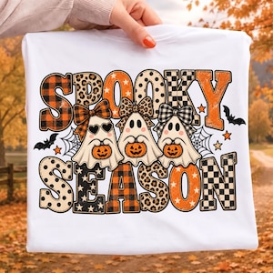 Op de afbeelding: Een wit t-shirt met de tekst SPOOKY SEASON in grote, gedessineerde letters. Het ontwerp toont drie geesten met strikjes die elk een kleine oranje pompoen vasthouden. De afbeelding is versierd met zwarte vleermuizen, spinnenwebben en oranje sterren.