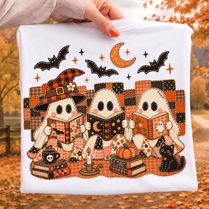 May include: A white t-shirt featuring a graphic of three ghosts reading books while sitting on a patchwork quilt. The design includes a black cat, a pumpkin, a candle, bats, and a crescent moon, all rendered in orange, black, and white tones.