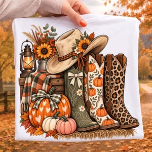 Può includere: Una t-shirt bianca con una stampa grafica di un cappello da cowboy beige decorato con girasoli, tre stivali western in verde, con motivo a zucca e stampa leopardata, una lanterna, una coperta a quadri e varie zucche.