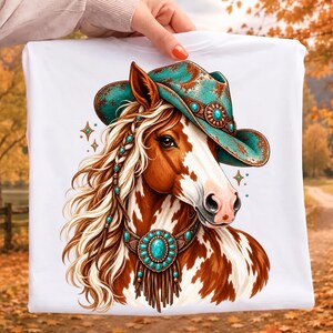May include: A white t-shirt featuring a graphic illustration of a brown and white pinto horse wearing a distressed turquoise cowboy hat and a matching turquoise gemstone collar with fringe accents. The horse has a braided mane with turquoise beads.