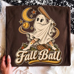May include: A dark brown t-shirt featuring a graphic of a white ghost wearing striped socks and holding a wooden baseball bat. The design includes a yellow crescent moon, stars, a baseball, a leather glove, and the text Fall Ball in a retro font.