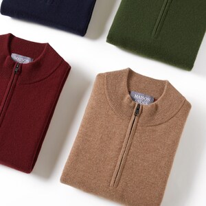Peut inclure: Quatre pulls pliés dans différentes couleurs : bleu marine, vert forêt, bordeaux et beige. Chaque pull a une fermeture éclair et une petite étiquette avec le texte "MAISON ANDES".