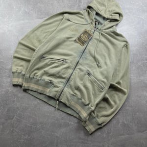 May include: A sage green zip-up hoodie with a hood and two front pockets. The hoodie has a tag attached and features a ribbed hem and cuffs with a cream stripe detail. The brand name is embroidered on the chest.