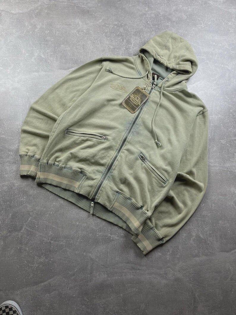 May include: A sage green zip-up hoodie with a hood and two front pockets. The hoodie has a tag attached and features a ribbed hem and cuffs with a cream stripe detail. The brand name is embroidered on the chest.