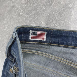 May include: A pair of blue denim jeans featuring a rectangular fabric label on the interior waistband. The label displays a small, embroidered American flag with red, white, and blue stripes and a blue field with white stars. The jeans have gold-tone metal rivets and visible tan stitching along the seams.
