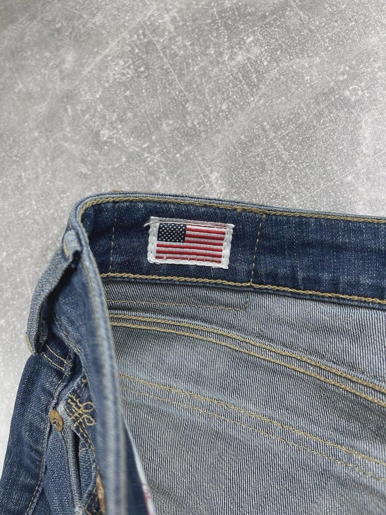 May include: A pair of blue denim jeans featuring a rectangular fabric label on the interior waistband. The label displays a small, embroidered American flag with red, white, and blue stripes and a blue field with white stars. The jeans have gold-tone metal rivets and visible tan stitching along the seams.