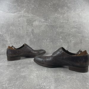 May include: A pair of dark brown leather dress shoes. The shoes have a low heel and a slip-on design. The leather has a textured appearance. The shoes are displayed on a gray surface.
