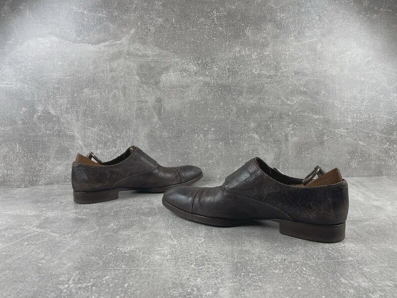 May include: A pair of dark brown leather dress shoes. The shoes have a low heel and a slip-on design. The leather has a textured appearance. The shoes are displayed on a gray surface.