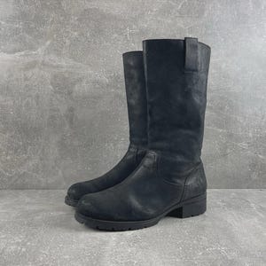 Könnte beinhalten: Ein Paar wadenhohe Stiefel aus mattem schwarzem Leder mit abgerundeter Zehenpartie, niedrigem Blockabsatz und einer vertikalen Zuglasche am Schaft. Die Stiefel haben sichtbare tonale Nähte am Vorderblatt und an der Fersenkappe sowie eine profilierte Sohle.