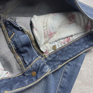 May include: Blue denim jeans featuring a visible pocket lining with a repeating red graphic print of a guitar-playing figure. The jeans include gold-toned metal rivets and thick, contrasting yellow stitching along the seams and waistband.