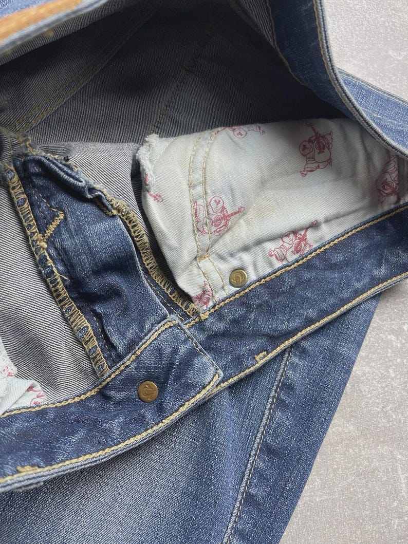 May include: Blue denim jeans featuring a visible pocket lining with a repeating red graphic print of a guitar-playing figure. The jeans include gold-toned metal rivets and thick, contrasting yellow stitching along the seams and waistband.