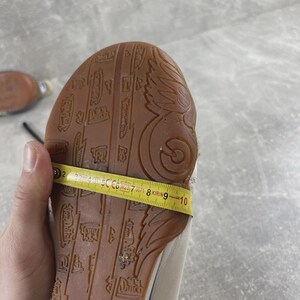May include: A tan shoe sole with a brown rubber tread featuring embossed Von Dutch logos and wing designs. A yellow measuring tape is wrapped across the width of the sole, showing a measurement of approximately 3.5 inches at the widest point.