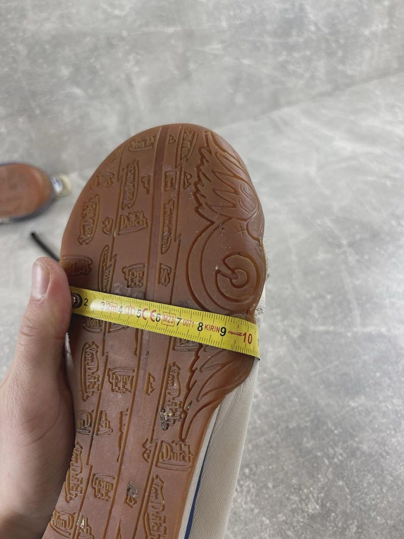 May include: A tan shoe sole with a brown rubber tread featuring embossed Von Dutch logos and wing designs. A yellow measuring tape is wrapped across the width of the sole, showing a measurement of approximately 3.5 inches at the widest point.