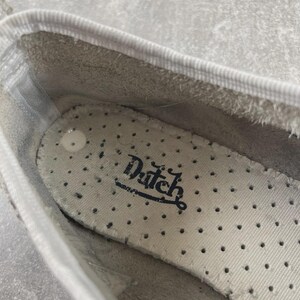 May include: The interior of a white shoe featuring a perforated off-white insole with the word Dutch printed in black script. The shoe has a white fabric trim along the top edge and a light gray textured lining visible inside the heel area.