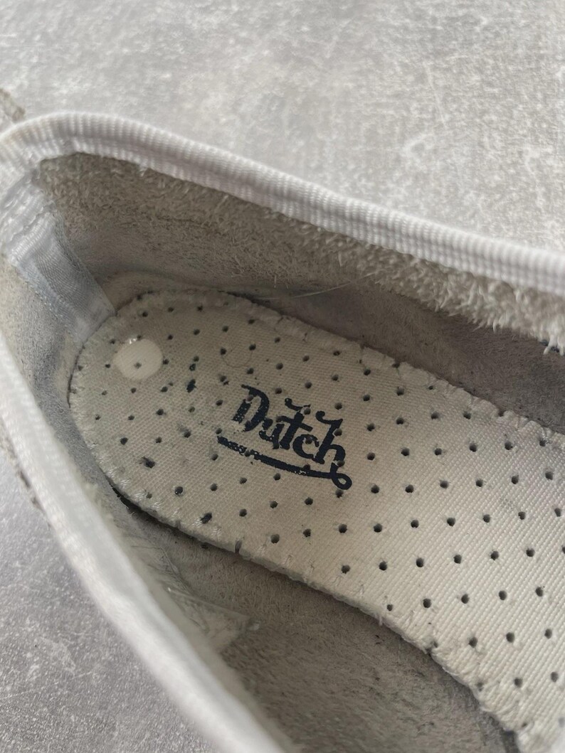 May include: The interior of a white shoe featuring a perforated off-white insole with the word Dutch printed in black script. The shoe has a white fabric trim along the top edge and a light gray textured lining visible inside the heel area.