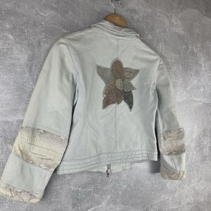 Peut inclure: Veste en cuir bleu clair avec un motif floral au dos. La veste est dot&eacute;e d'une fermeture &eacute;clair et de patchs d&eacute;coratifs sur les manches. Le motif floral est compos&eacute; de nuances de marron et de vert.