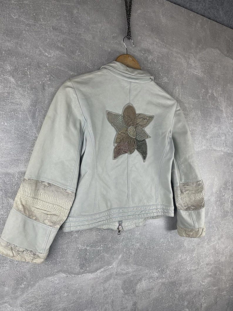 Peut inclure: Veste en cuir bleu clair avec un motif floral au dos. La veste est dot&eacute;e d'une fermeture &eacute;clair et de patchs d&eacute;coratifs sur les manches. Le motif floral est compos&eacute; de nuances de marron et de vert.